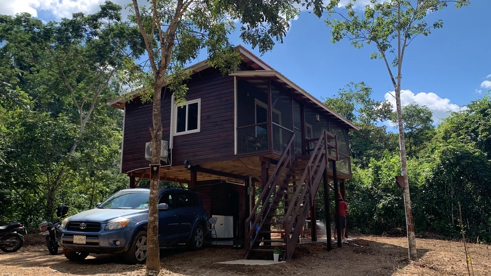 The cabin stained dark, with a car and motorbike parked beneath it under a blue sky