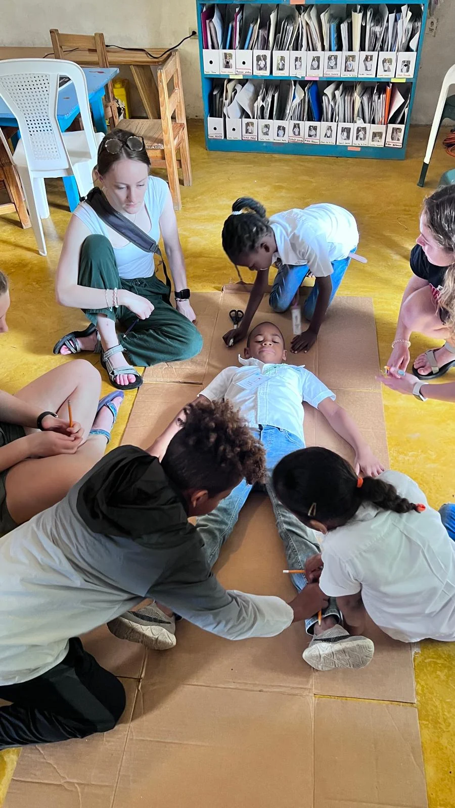 Volunteers and local children kneeling on cardboard to trace the outline of a boy lying down during a classroom activity